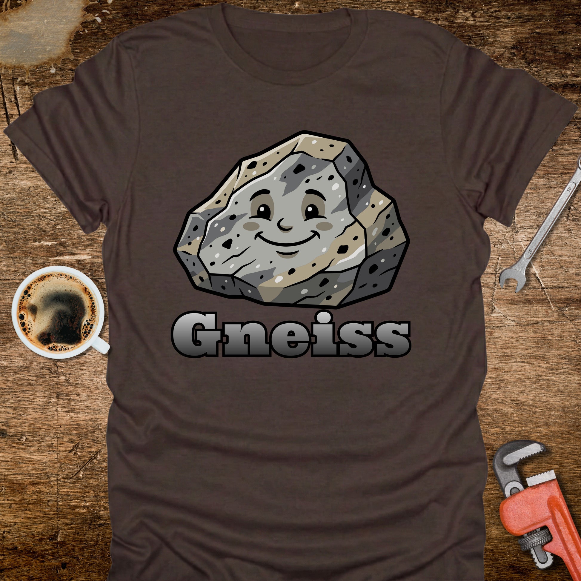 Brown t-shirt with a smiling rock graphic and 'Gneiss' text on a wooden surface with coffee and tools.