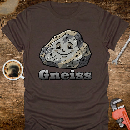 Brown t-shirt with a smiling rock graphic and 'Gneiss' text on a wooden surface with coffee and tools.