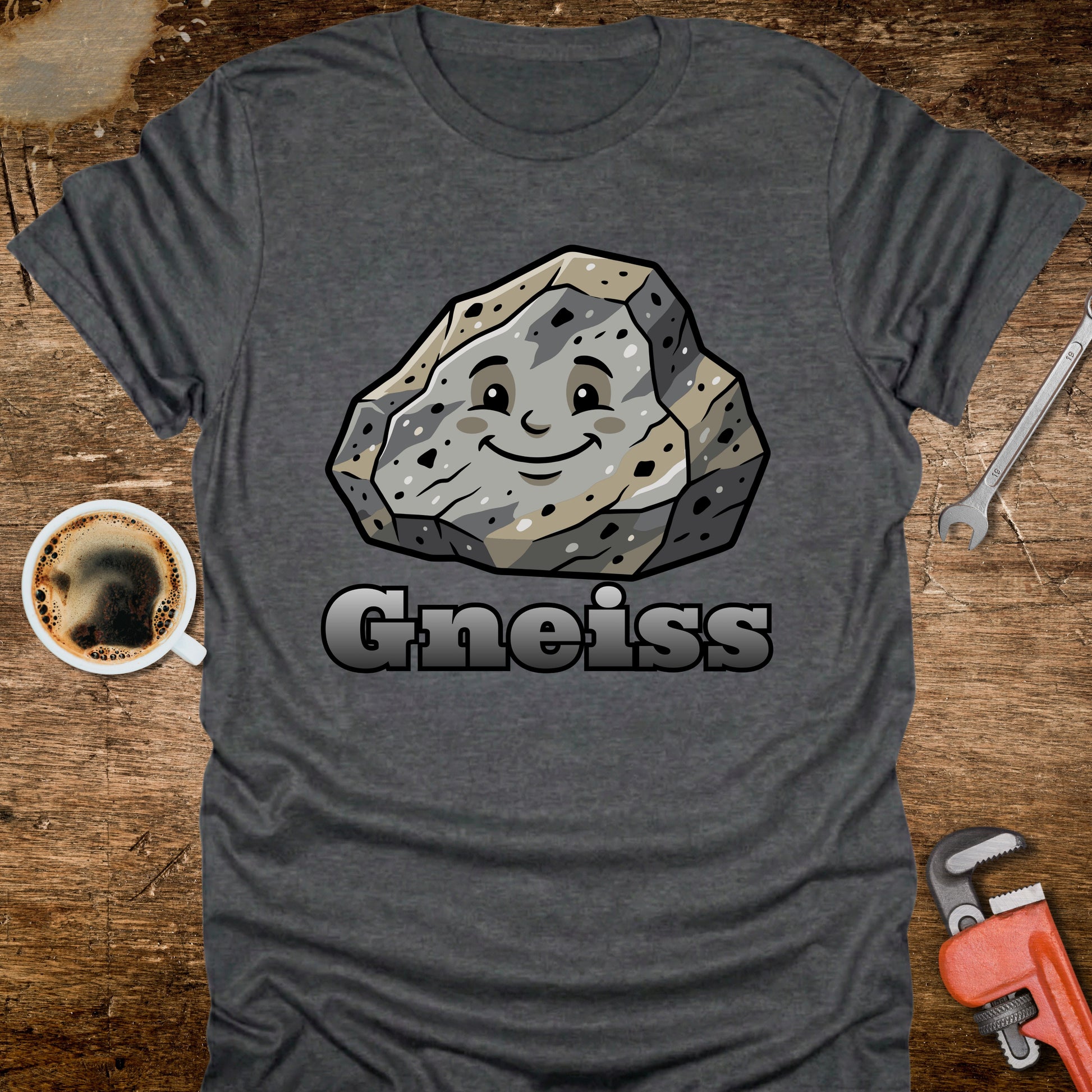 Gray t-shirt with a cartoon gneiss stone graphic and the word 'Gneiss' on a wooden surface with a coffee cup and tools.
