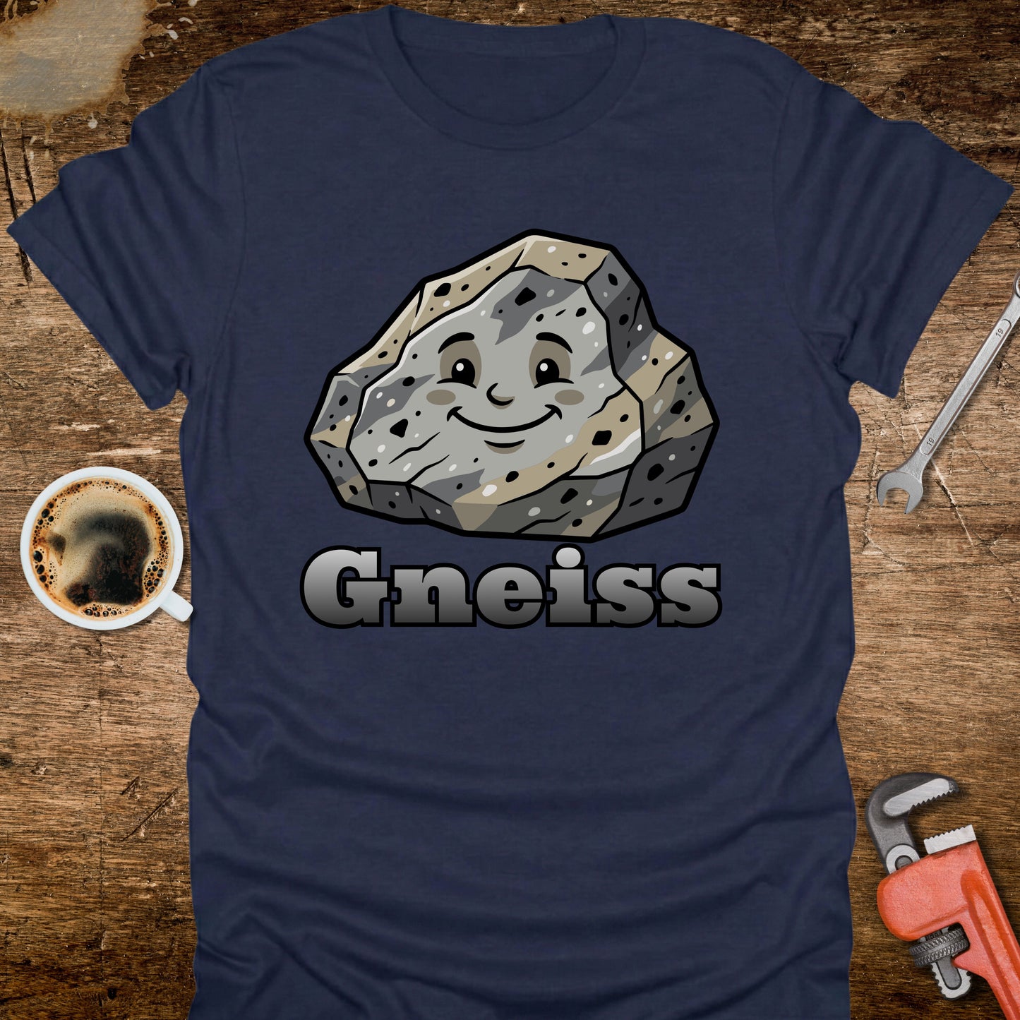 Navy blue t-shirt with a cartoon gneiss stone graphic and 'Gneiss' text on a wooden surface with tools and coffee.
