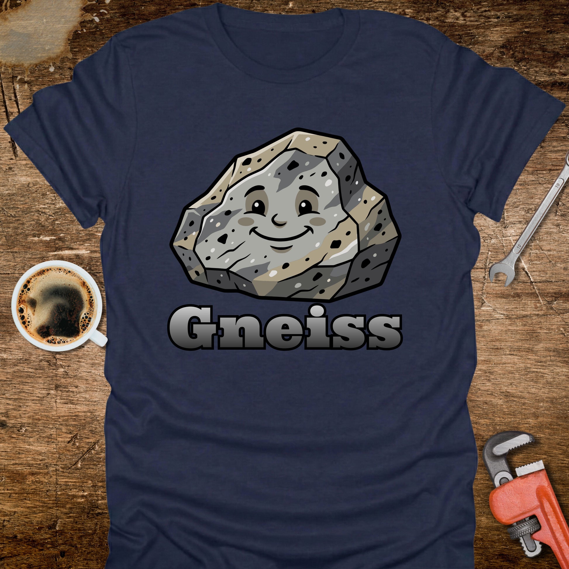 Navy blue t-shirt with a cartoon gneiss stone graphic and 'Gneiss' text on a wooden surface with tools and coffee.