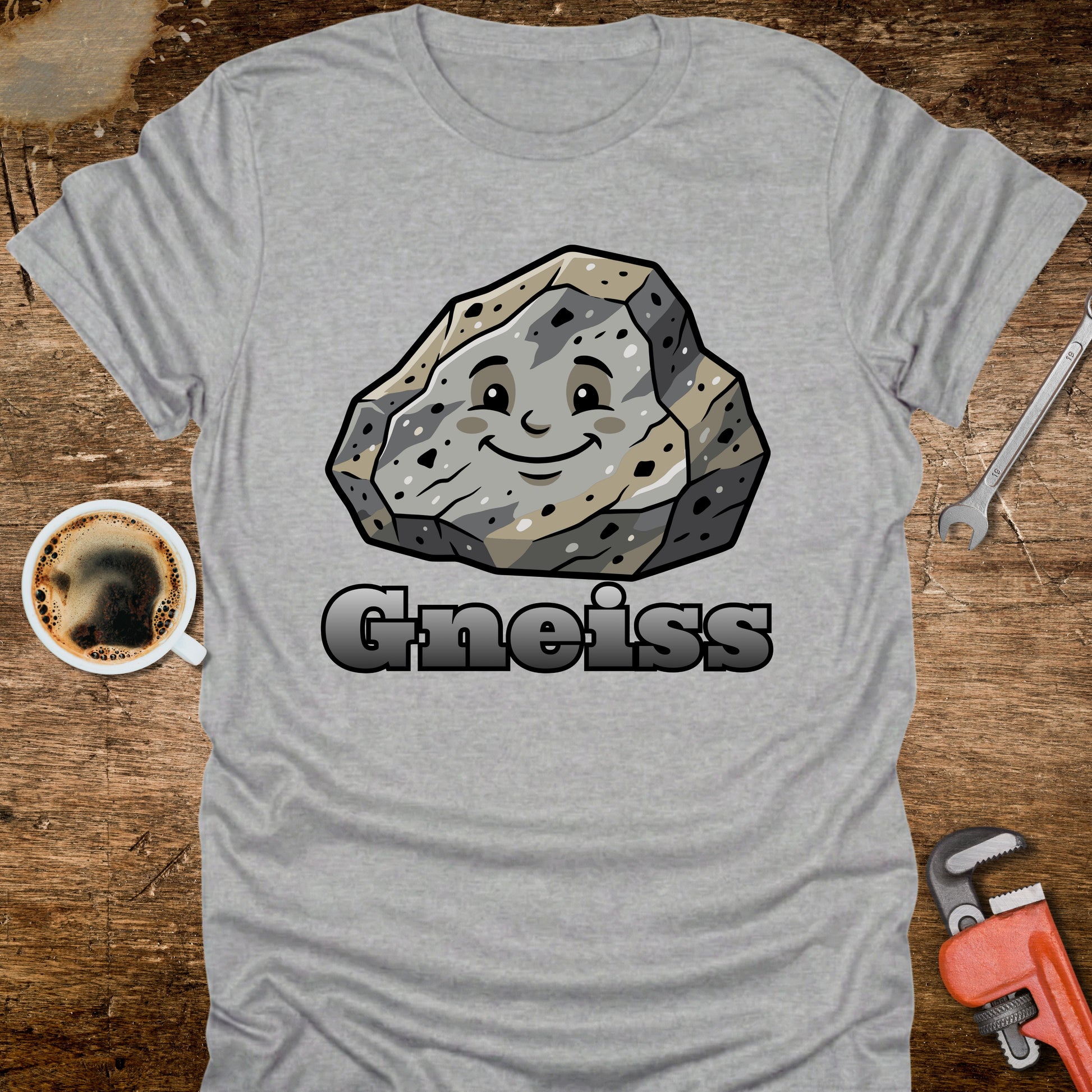 Gray t-shirt with a cartoon gneiss stone graphic and the word 'Gneiss' on a wooden surface with a coffee cup and tools.