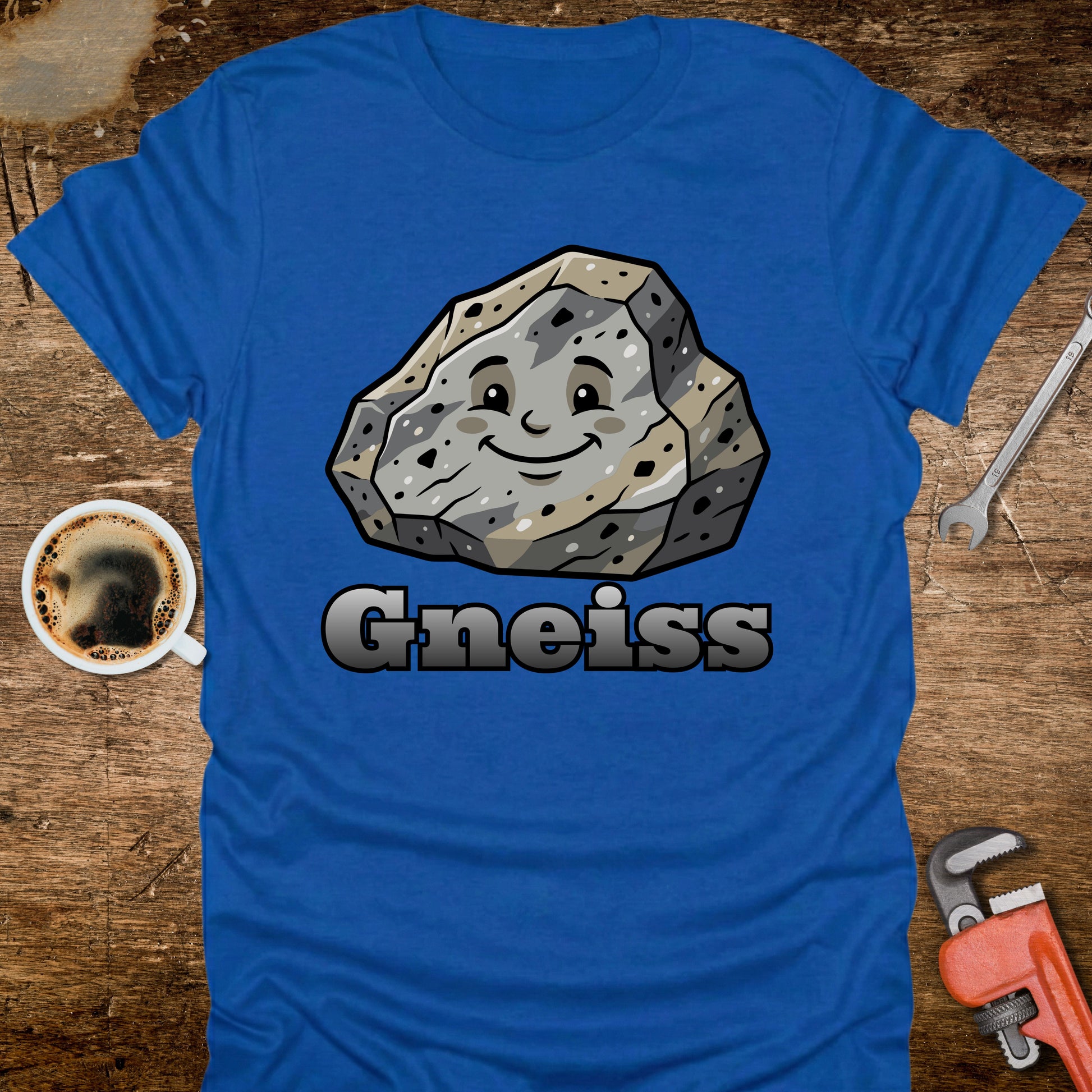 Blue t-shirt with a smiling gneiss rock graphic and 'Gneiss' text on a wooden surface with tools and a coffee cup.