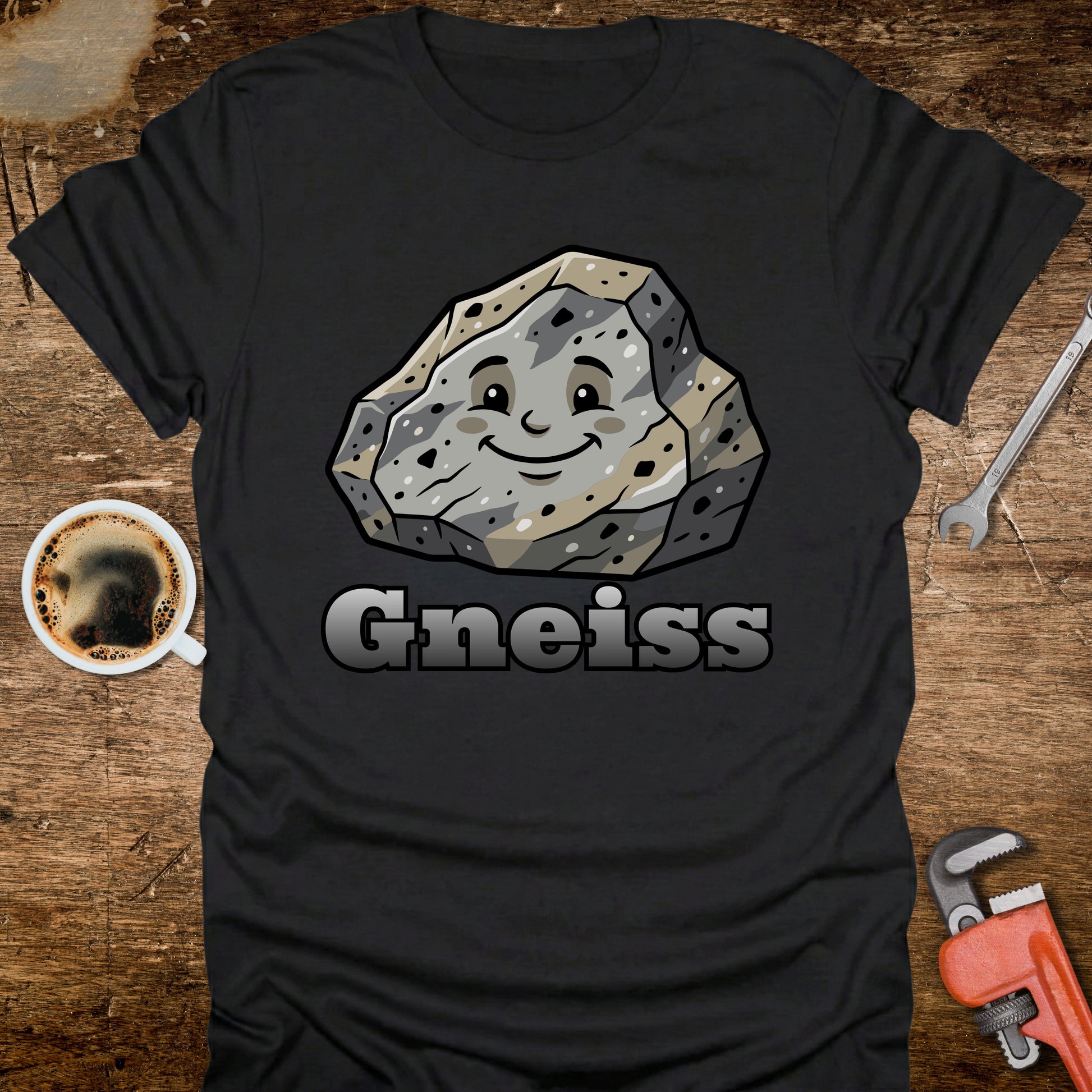 Black t-shirt with a smiling gneiss stone graphic and 'Gneiss' text on a wooden surface with tools and a coffee cup.