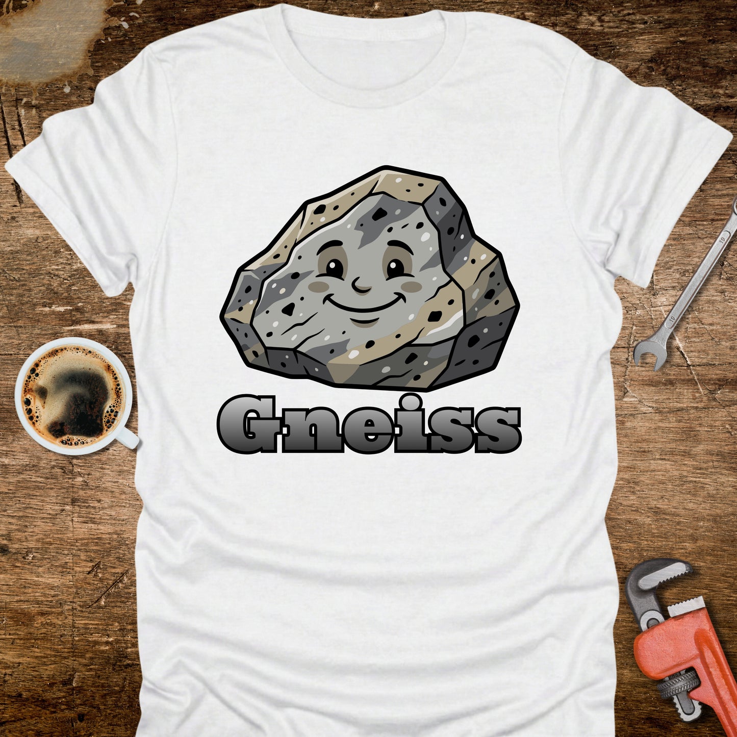 White t-shirt with a cartoon gneiss rock graphic and 'Gneiss' text on a wooden surface with tools and a coffee cup.