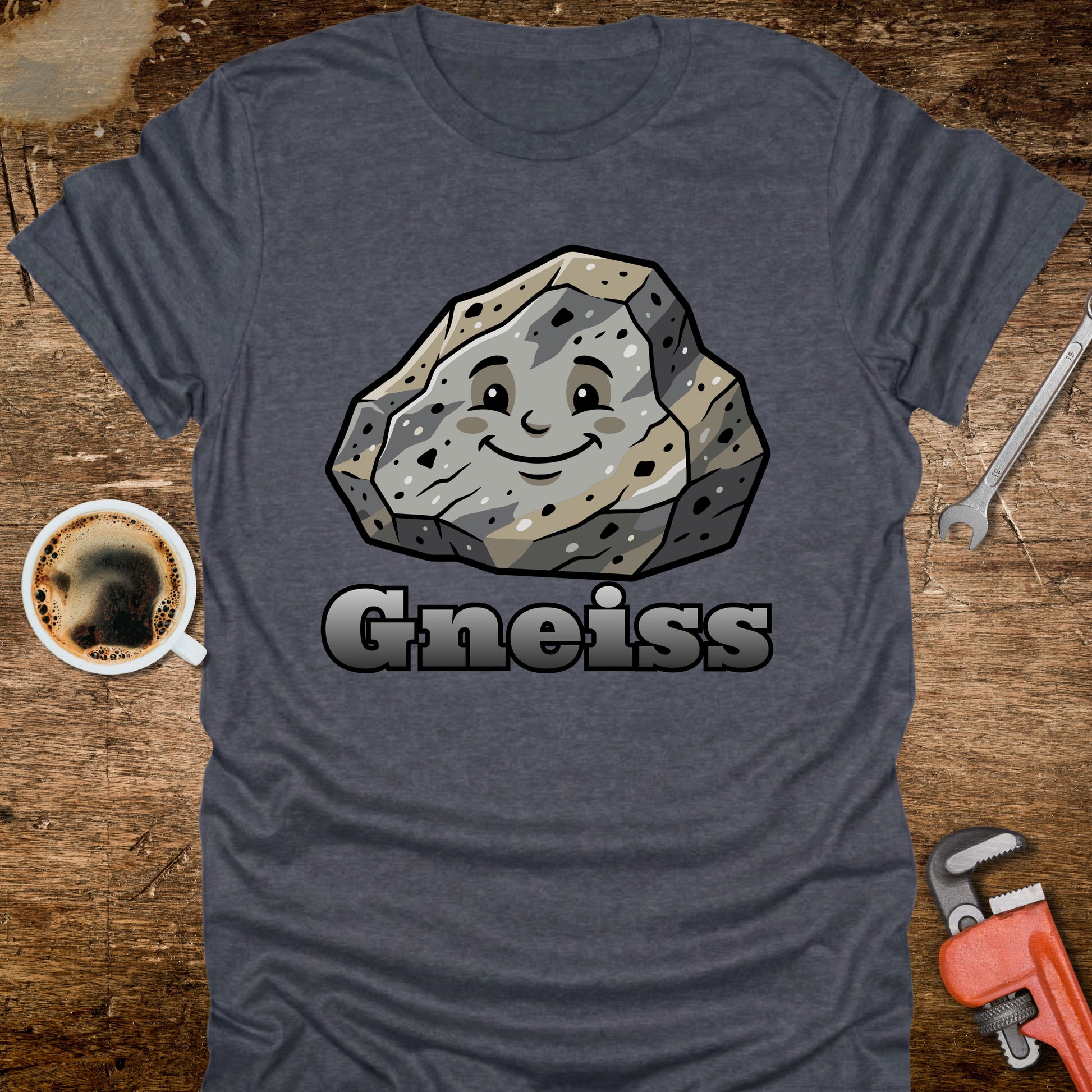 Blue t-shirt with a cartoon gneiss stone graphic and the word 'Gneiss' on a wooden surface with a coffee cup and tools.