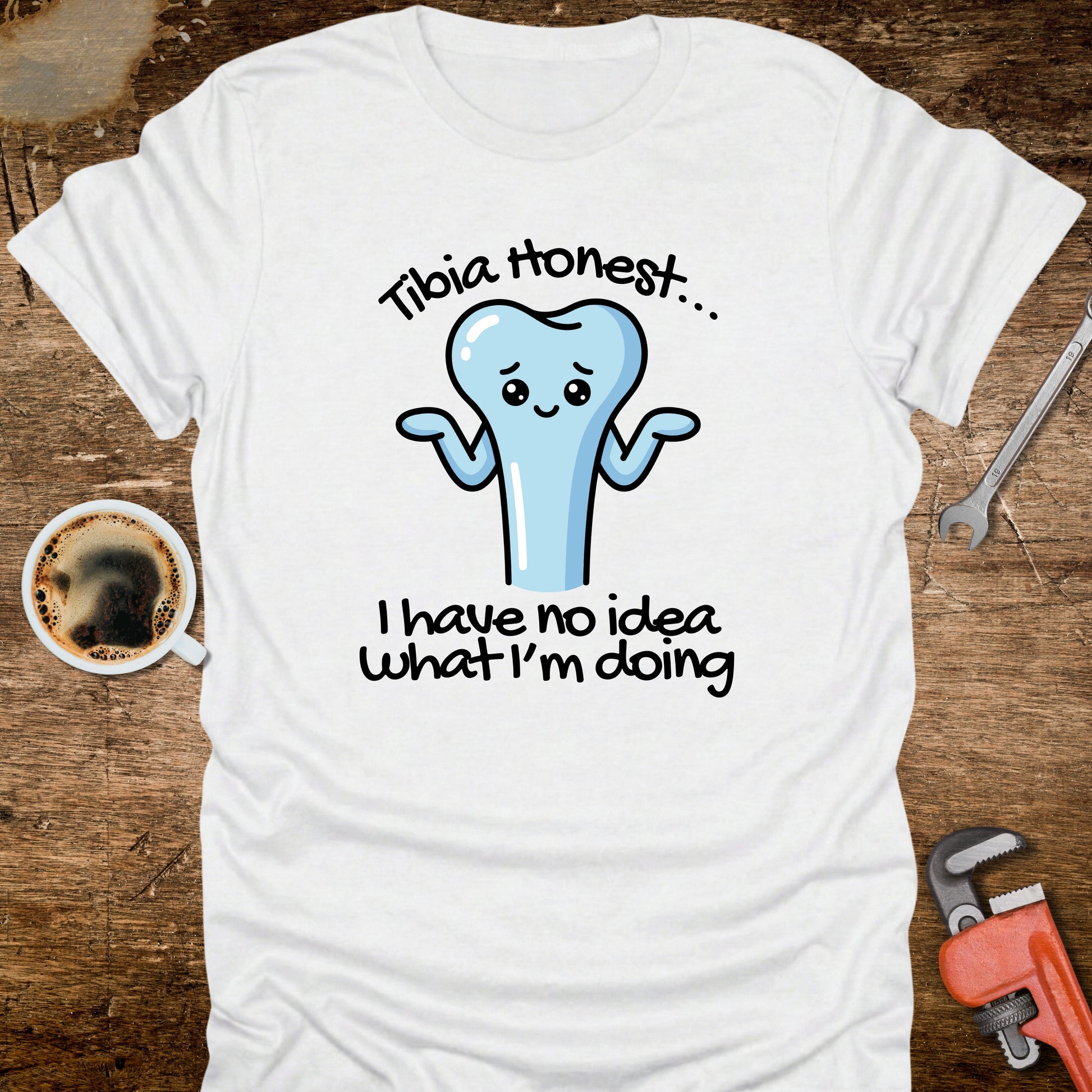 White t-shirt with a cartoon tooth graphic and text on a wooden surface with coffee and tools.