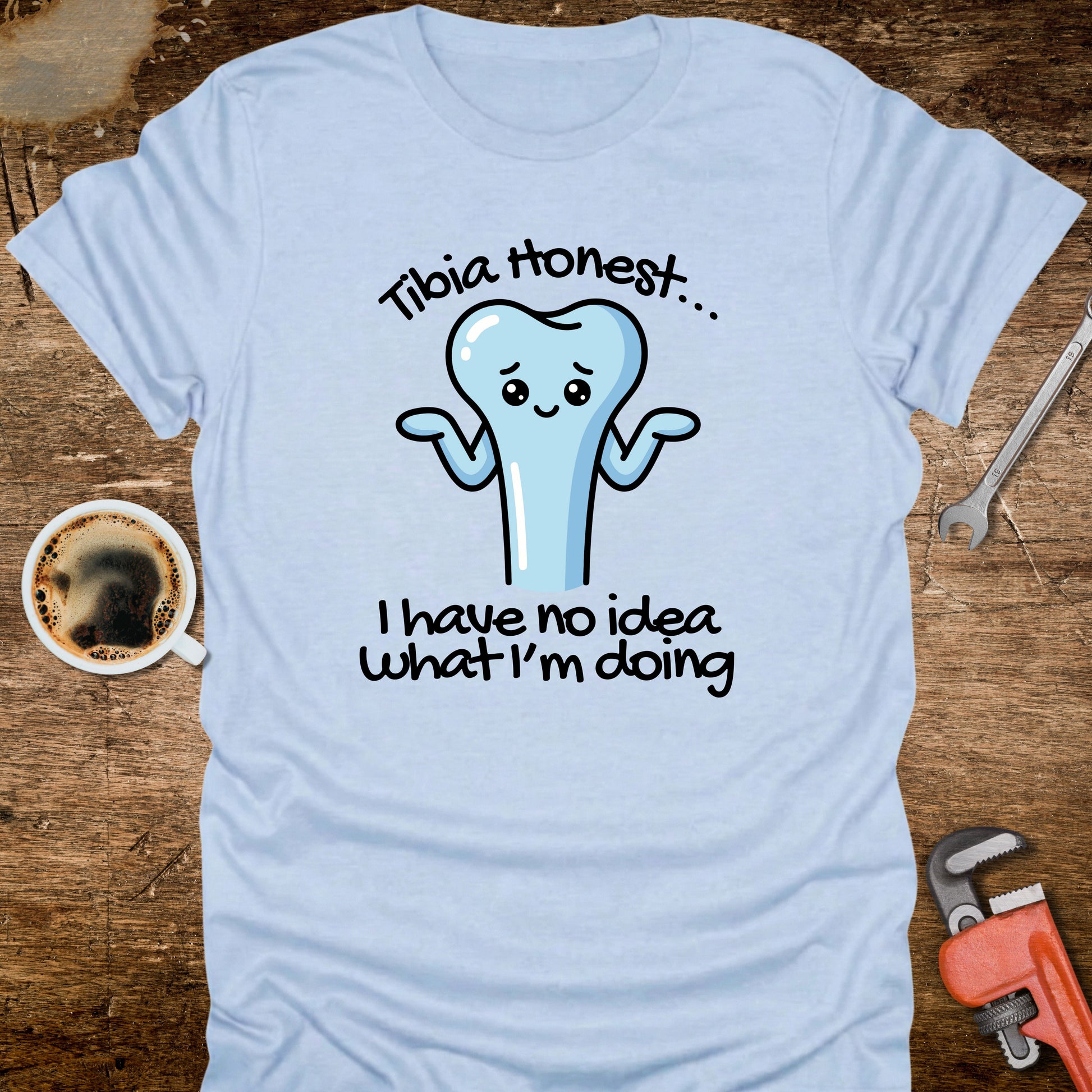 Light blue t-shirt with a cartoon tooth graphic and text on a wooden surface with tools and a coffee cup.