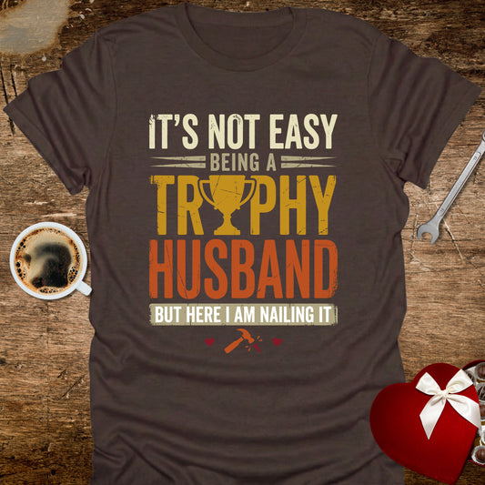 Brown t-shirt with humorous text design on a wooden surface with coffee and heart-shaped gift box.